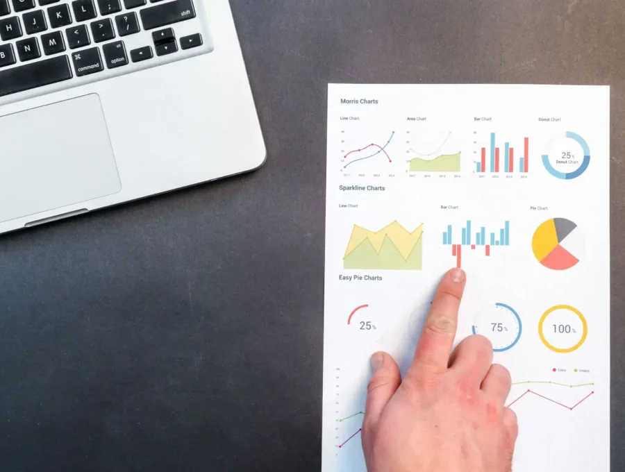 A business professional pointing at a printed analytics dashboard displaying charts and performance metrics beside a laptop on a desk.