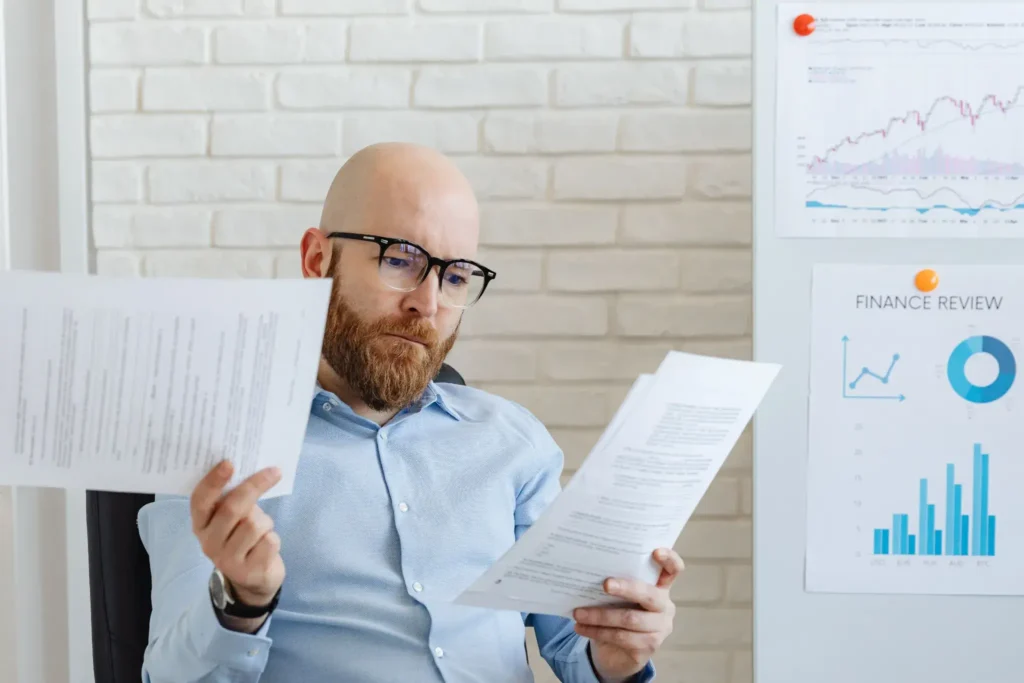 Business professional reviewing printed reports in an office with financial charts displayed on the wall.